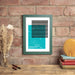 A framed lyric print hangs on a brick wall above a wooden shelf with books, a vase of dried flowers, and decorative items. The print features a turquoise block with black horizontal lyric strips and white text below that reads: “Outnumbered, Dermot Kennedy – Kindreds Meet Again – October 22, 2022.” The frame is green and modern in style