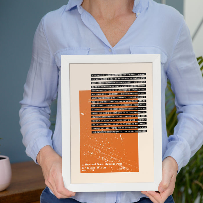 A person in a light blue shirt holds a framed lyric print with an orange textured block and black strip text. The song lyrics are from “A Thousand Years” by Christina Perri. The bottom section reads: “A Thousand Years, Christina Perri – Mr & Mrs Wilson – Dec 12, 2021.” The design has a clean, modern feel with a white frame and beige background.