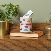 Ceramic money jar labeled ‘Las Vegas Fund’ with an illustrated Las Vegas skyline, shown with a £5 note inserted, displayed on a book with plants and décor in the background.