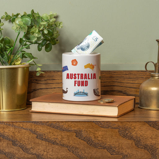 Ceramic money jar labeled ‘Las Vegas Fund’ with an illustrated Las Vegas skyline, shown with a £5 note inserted, displayed on a book with plants and décor in the background.