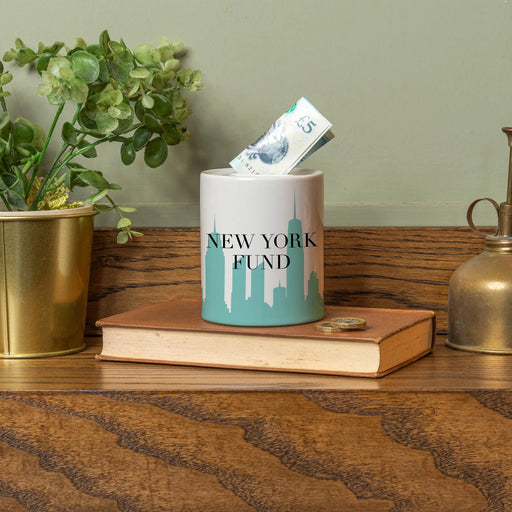 Ceramic money jar with New York City skyline and the words ‘New York Fund,’ sitting on a book with a £5 note sticking out, styled with plants and decorative items in the background.”