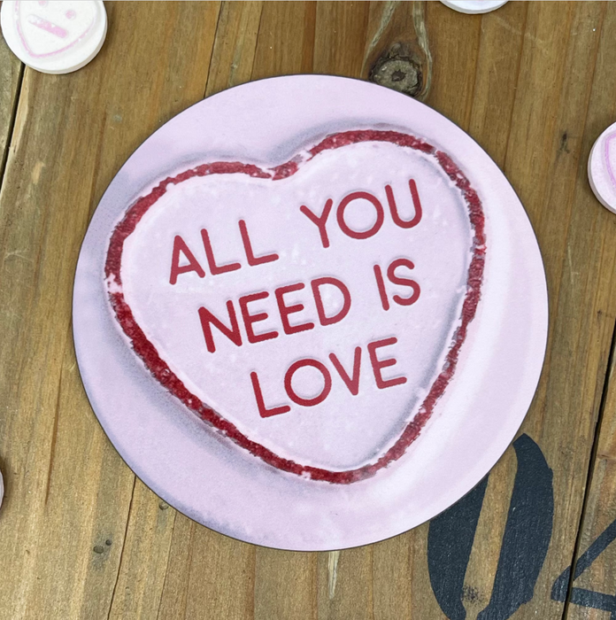 Close-up of pink love heart coaster with the phrase “All You Need Is Love,” styled on a rustic wooden background with small candy hearts.