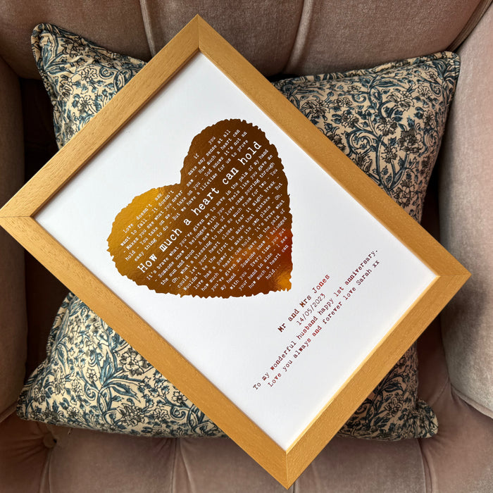 Framed gold foil heart print reading “How much a heart can hold,” personalised with couple’s names and anniversary date, resting on a patterned cushion.