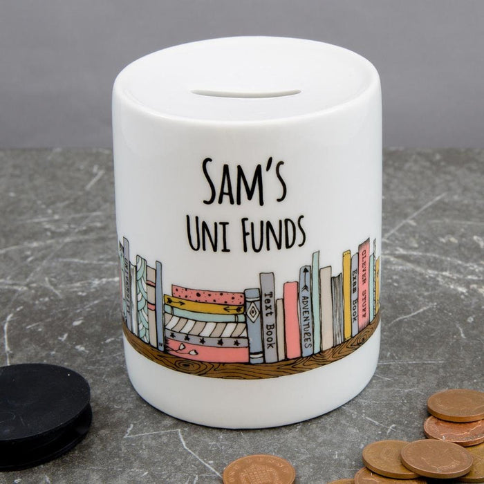 Ceramic money box with illustration of colourful books and the text “Sam’s Uni Funds,” placed on a grey surface with books and coins around.
