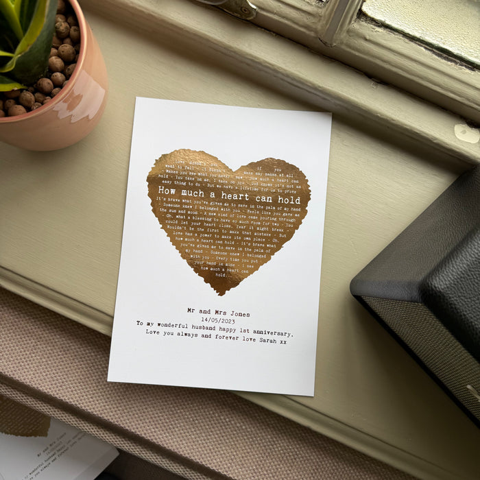 Gold foil heart lyric print with the words “How much a heart can hold,” personalised with names and anniversary message, displayed on a windowsill beside a plant pot.