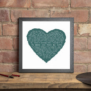 Two framed heart-shaped lyric prints displayed against a neutral gray wall. The larger print, in a black frame, features a pink heart with song lyrics and the message “Happy Anniversary – To my beautiful wife of 24 years, all my love, Peter x.” The smaller print, in a light oak frame, shows a blue heart with lyrics centered around the line “I wanna stay with you until we’re grey and old,” and is personalized with “Mr & Mrs Luca – Bardale Hall – 25 March 2017.” Both prints have a clean white background and a