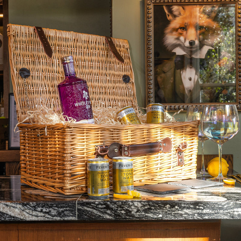 Luxury wicker gin hamper containing Whitley Neill gin, Fever-Tree tonic cans, and straw filler, styled on a bar countertop with glasses and lemons.