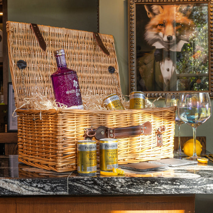 Luxury wicker gin hamper containing Whitley Neill gin, Fever-Tree tonic cans, and straw filler, styled on a bar countertop with glasses and lemons.