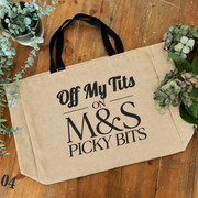 A natural beige canvas tote bag with black handles, printed with the humorous text 'Off My Tits ON M&S PICKY BITS' in black lettering, displayed on a rustic wooden tabletop with decorative green plants.