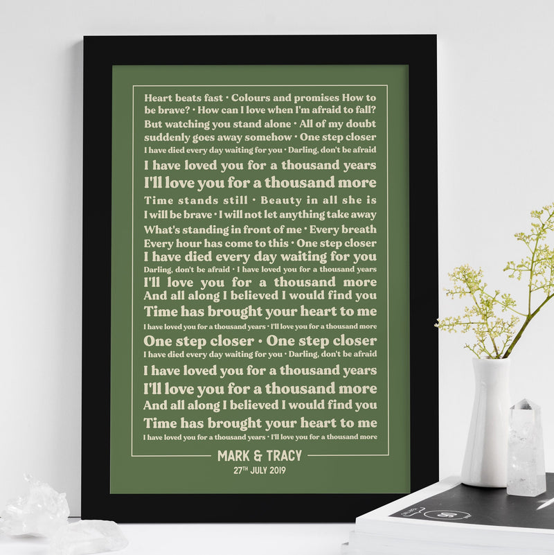 A green lyric print framed in black, standing on a white tabletop with a vase and a small plant beside it. The text is in cream-colored font and features the lyrics to “A Thousand Years” by Christina Perri. Prominent lines include “I’ll love you for a thousand years, I’ll love you for a thousand more.” The personalization at the bottom reads “MARK & TRACY – 27TH JULY 2019,” making it a timeless anniversary keepsake.