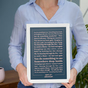 A person wearing a light blue shirt holds a framed lyric print with a white frame and dark blue background. The text features the lyrics to “You Do Something to Me” in cream-colored type, centered in neat alignment. The words “HAPPY 25TH ANNIVERSARY” are printed at the bottom. The minimalist design and muted tones give the print an elegant, personalized anniversary feel.