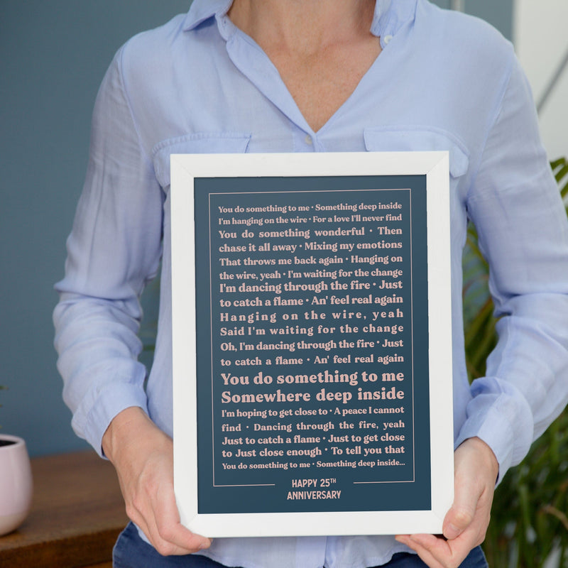A person wearing a light blue shirt holds a framed lyric print with a white frame and dark blue background. The text features the lyrics to “You Do Something to Me” in cream-colored type, centered in neat alignment. The words “HAPPY 25TH ANNIVERSARY” are printed at the bottom. The minimalist design and muted tones give the print an elegant, personalized anniversary feel.