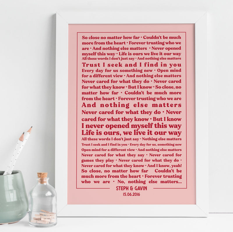 A framed lyric print with a white frame and dusty rose background, displayed on a white desk with a small vase, pencil pot, and decorative bottle. The text showcases lyrics from “Nothing Else Matters” in red type, with emphasis on lines such as “Trust I seek and I find in you” and “Life is ours, we live it our way.” The personalization at the bottom reads “STEPH & GAVIN – 15.06.2016,” giving the artwork a romantic, modern look.