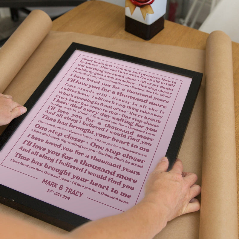 A close-up of a framed lyric print being wrapped in brown paper. The print features the same “A Thousand Years” lyrics as the previous image, but with a pink background and dark red text inside a black frame. The names “MARK & TRACY – 27TH JULY 2019” appear at the bottom. The scene shows hands gently packaging the print, emphasizing its role as a thoughtful, personalized gift.