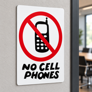 A rectangular white sign mounted on a light gray wall, featuring a red prohibition symbol over a black mobile phone icon and the words 'NO CELL PHONES' in black. A blurred office interior is visible in the background.