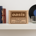 Oasis engraved wooden sign displayed on a white shelf beside books and a vinyl record, creating a modern music-themed home decor se