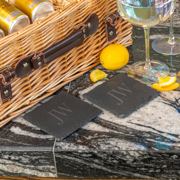 Pair of personalised slate coasters engraved with “JW,” displayed next to a wicker hamper, lemons, and wine glasses on a marble countertop.