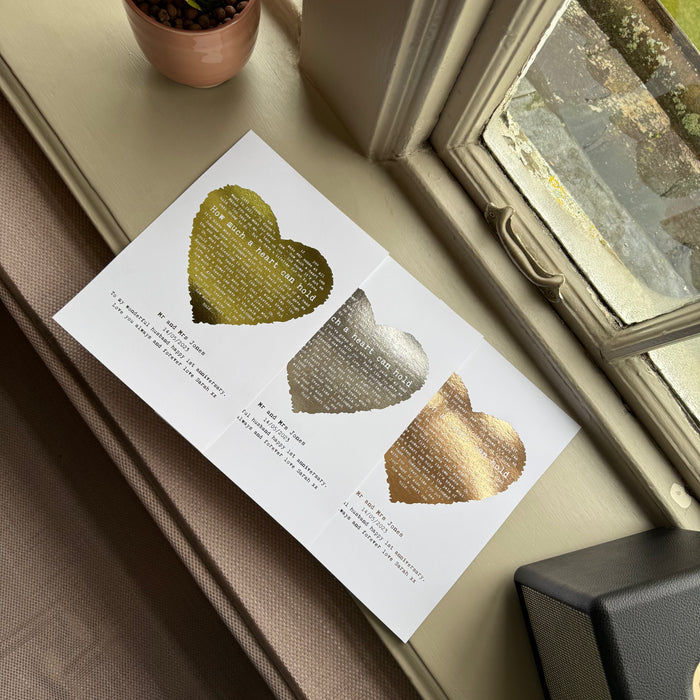 Personalised foil heart lyric prints in gold, silver, and rose gold finishes, showing custom names and dates, laid out on a windowsill beside a plant.
