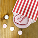 Personalised pink love heart coaster with names “Vicky & Hamish” partially tucked into a red and white striped paper bag, surrounded by pastel sweets.