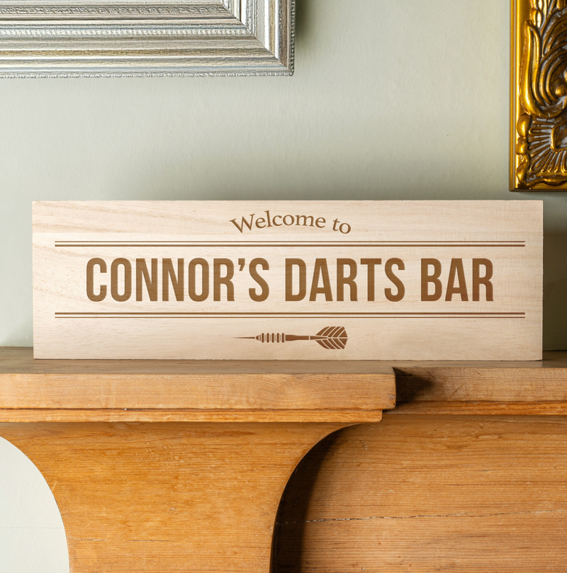 Personalised wooden darts bar sign engraved with “Connor’s Darts Bar”, displayed on a wooden mantelpiece.