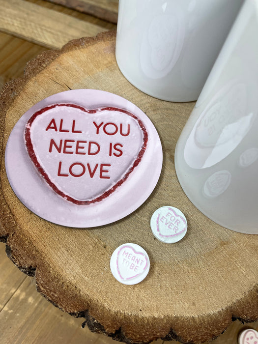 Pink “All You Need Is Love” coaster placed on a wooden log slice with small heart sweets and a mug nearby, styled for a cosy romantic setting.