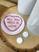 Pink “All You Need Is Love” coaster placed on a wooden log slice with small heart sweets and a mug nearby, styled for a cosy romantic setting.