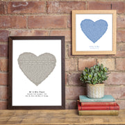 Two framed lyric prints displayed on a rustic brick wall. The larger print, in a dark wood frame, features a taupe heart with lyrics from “Simply the Best” and the inscription “Mr & Mrs Finch – Saturday 30th December 2022 – Take My Heart and Make it Strong.” The smaller print, in a light oak frame, shows a blue heart with lyrics from “All of Me” and the text “Katie & May – 1st March 2010.” Both prints are accompanied by a small green plant and stacked vintage books.