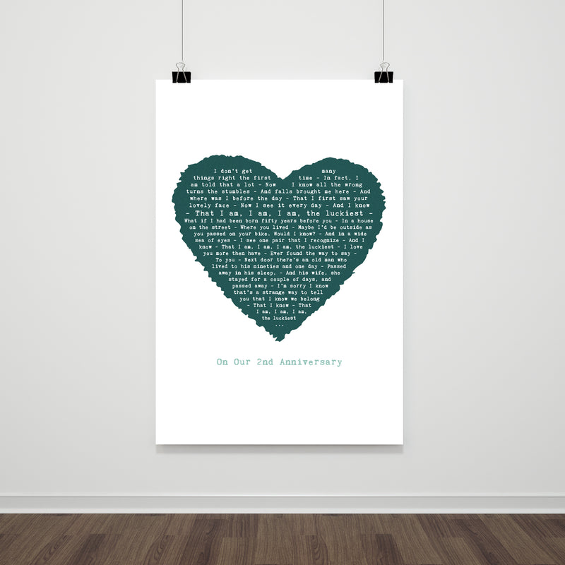 A large lyric print hanging on a white wall, featuring a deep teal heart composed of the words from “The Luckiest.” Below the heart, the message reads “On Our 2nd Anniversary.” The clean, uncluttered layout and hanging clips give it a modern and minimalist appearance.