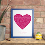 A lyric print framed in bold blue, showing a deep magenta heart filled with the words of “Lucky.” The personalization reads “Mr and Mrs Bedford – Love Sue xx.” The artwork is displayed on a rustic wooden surface with a yellow patterned plant pot and brick wall backdrop, giving it a cozy, heartfelt charm.
