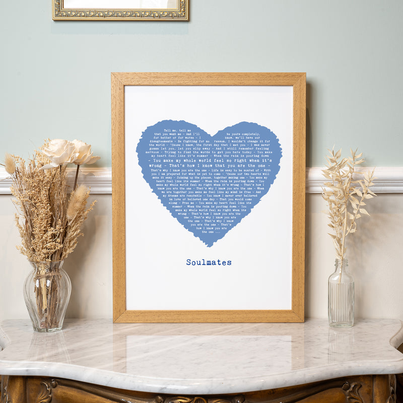 A framed lyric art print with a blue heart containing song lyrics, centered above the word “Soulmates.” The artwork is displayed on a marble-topped table with dried flowers in glass vases on either side. The print has a light oak frame and sits against a pale green wall, evoking a calm and loving feel.