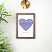 A framed lyric print with a lavender-purple heart featuring the lyrics of “God Only Knows.” The design is personalized with “Mum and Dad” beneath the heart and hangs on a light wall with a brass light fixture above and green plant leaves framing the scene. The frame is dark wood for a timeless look.