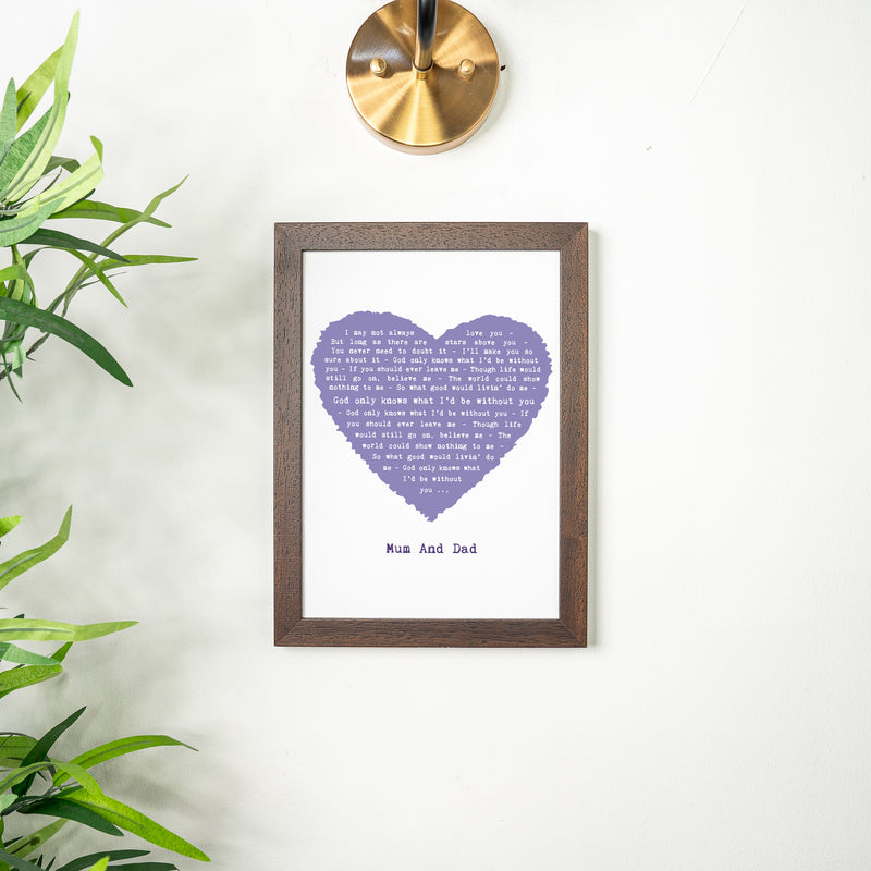 A framed lyric print with a lavender-purple heart featuring the lyrics of “God Only Knows.” The design is personalized with “Mum and Dad” beneath the heart and hangs on a light wall with a brass light fixture above and green plant leaves framing the scene. The frame is dark wood for a timeless look.