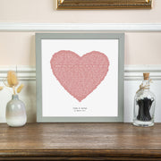 A framed lyric print with a dusty pink heart filled with song lyrics, displayed on a wooden surface against a soft cream wall. Below the heart, the personalization reads “John & Karen – 25 March 2017.” The print is in a light gray frame, surrounded by delicate dried flowers and decor, creating a warm and elegant atmosphere.