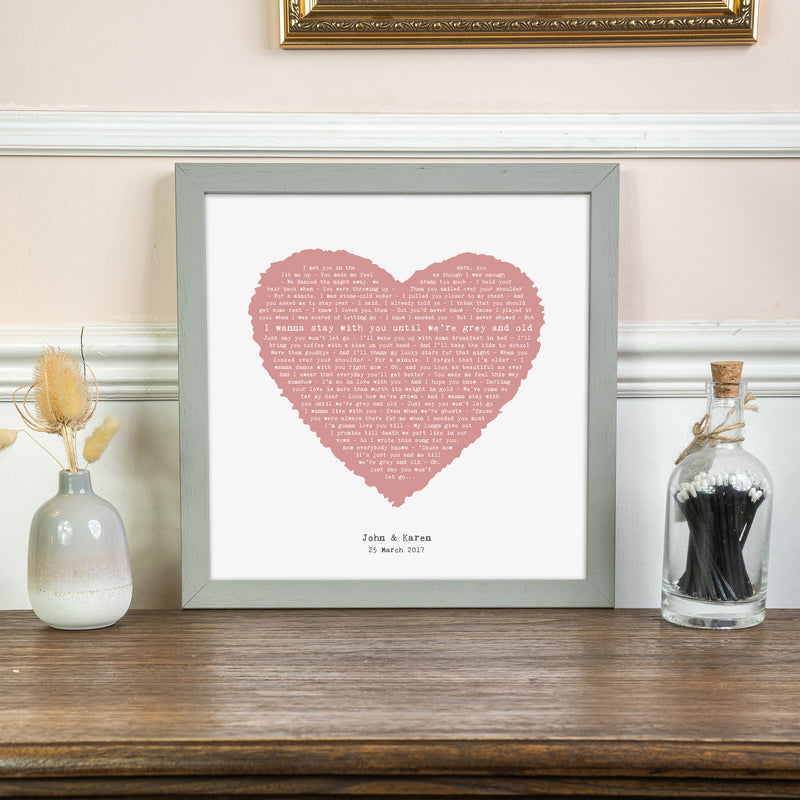A framed lyric print with a dusty pink heart filled with song lyrics, displayed on a wooden surface against a soft cream wall. Below the heart, the personalization reads “John & Karen – 25 March 2017.” The print is in a light gray frame, surrounded by delicate dried flowers and decor, creating a warm and elegant atmosphere.