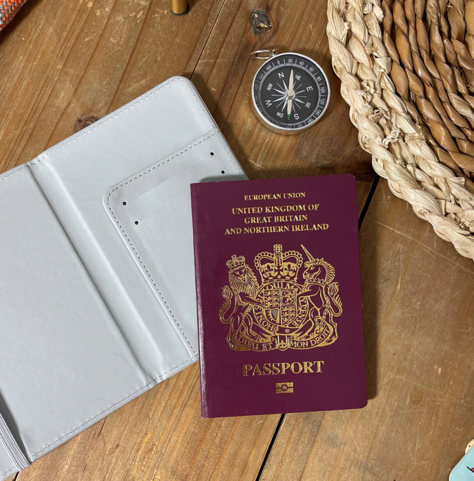 A travel-themed flat lay featuring a British passport, an open white faux leather passport holder, a compass, and a woven travel bag on a rustic wooden surface. The image highlights the inside of the passport cover with card pockets.