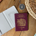 A travel-themed flat lay featuring a British passport, an open white faux leather passport holder, a compass, and a woven travel bag on a rustic wooden surface. The image highlights the inside of the passport cover with card pockets.