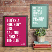 Two framed quote prints displayed on a rustic brick wall above a wooden shelf with books and a small potted plant. The larger print on the left has a deep pink background and bold white text reading: “You’re a pink pony girl, and you dance at the club.” The smaller print on the right has a mint green background with the words: “I’ve loved you for a thousand years, I’ll love you for a thousand more.” Both feature clean, modern typography.