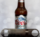 Textured gold metal bottle opener showing the reverse side, positioned in front of a Coors Light beer bottle on a grey surface.