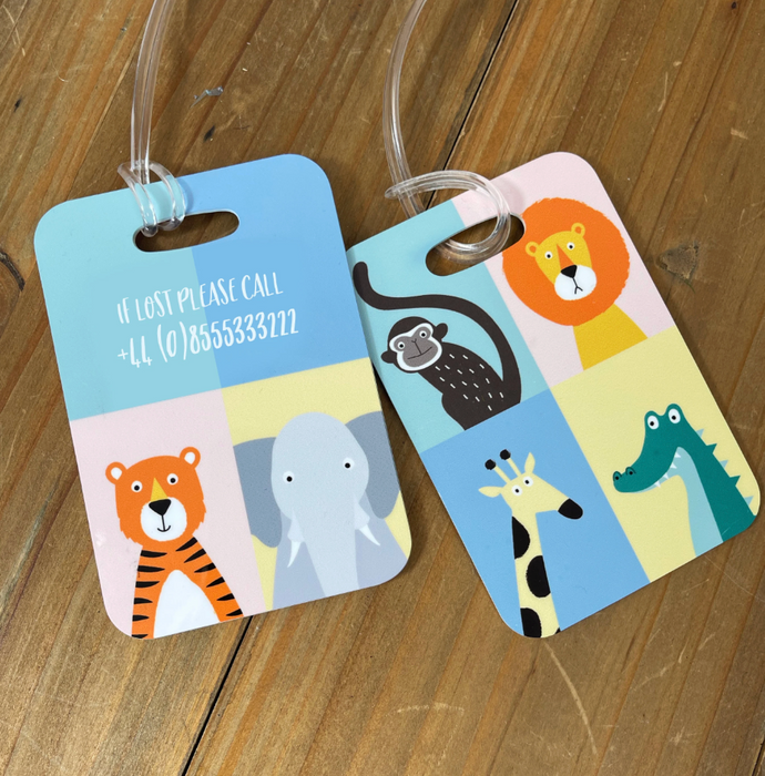 Two colourful children’s luggage tags featuring cute animal illustrations — a tiger, elephant, monkey, lion, giraffe, and crocodile — arranged in pastel squares. One tag includes a printed contact message reading “If lost please call +44 (0)8555333222.” Tags are displayed on a wooden table.