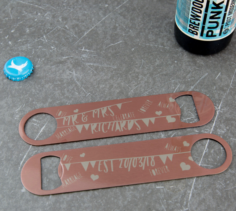 Two engraved bronze bar blade bottle openers with personalised wedding designs and BrewDog bottle cap, displayed on a grey textured surface.