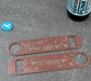 Two engraved bronze bar blade bottle openers with personalised wedding designs and BrewDog bottle cap, displayed on a grey textured surface.
