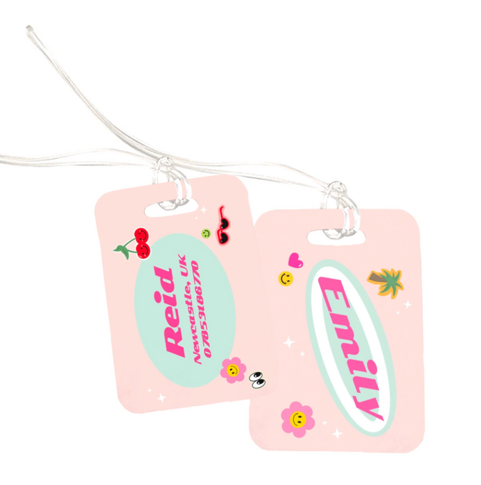 Two matching pink luggage tags with fun sticker-style graphics including cherries, hearts, flowers, sunglasses, and smiley faces. Each tag is personalised — one with the name Emily and the other with Reid, city, and contact details — attached to clear plastic loops for securing to luggage.