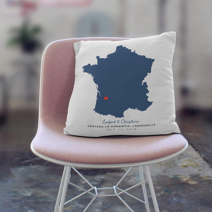 White cushion with navy blue map of France and red heart marking a location, personalised with names and wedding venue text, shown on a pale pink chair.