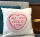 White cushion with pink heart design and the phrase “All You Need Is Love”, displayed on a grey chair with a teal cushion behind.