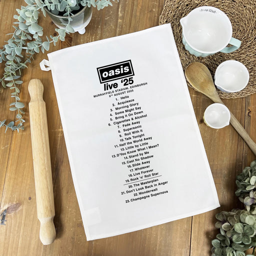 White tea towel printed with “Oasis Live ’25” concert setlist, styled on a wooden table with kitchen accessories including a rolling pin, wooden spoons, and ceramic bowls.