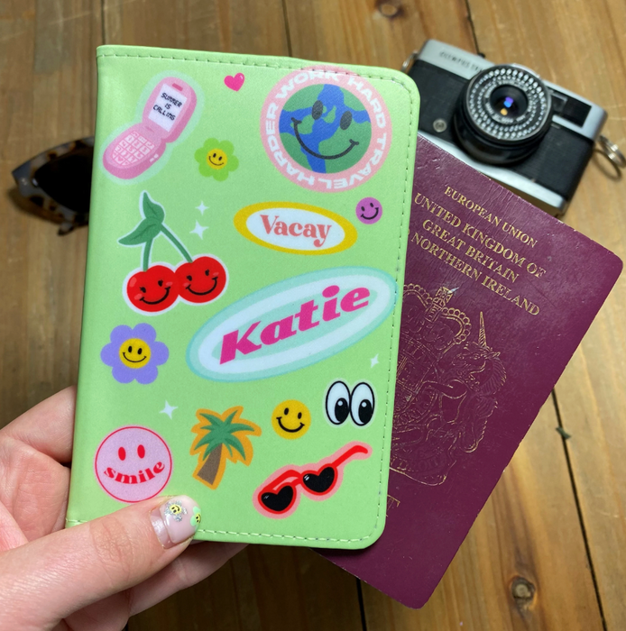A close-up of a lime green personalised passport holder decorated with vibrant stickers including cherries, a smiley flower, a palm tree, sunglasses, and a globe. The name Katie appears in pink and white retro lettering. The holder is displayed with a maroon passport and vintage camera on a wooden surface.
