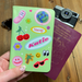 A close-up of a lime green personalised passport holder decorated with vibrant stickers including cherries, a smiley flower, a palm tree, sunglasses, and a globe. The name Katie appears in pink and white retro lettering. The holder is displayed with a maroon passport and vintage camera on a wooden surface.