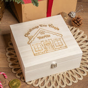 A beige-colored wooden Christmas Eve box with laser engravings, placed on a doily, with a rope handle, surrounded by festive decorations like pine cones and a red ribbon.