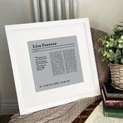 A framed lyric print of the song “Live Forever” by Oasis displayed on a cozy surface with books and a knitted blanket. The artwork features a gray background and black text with the lyric quote: “Maybe you’re the same as me, we see things they’ll never see. You and I are gonna live forever.” It’s personalized with “Dad – 65 years and still rocking it. Love Amy & Tom x.” The frame is white with a clean, modern design.