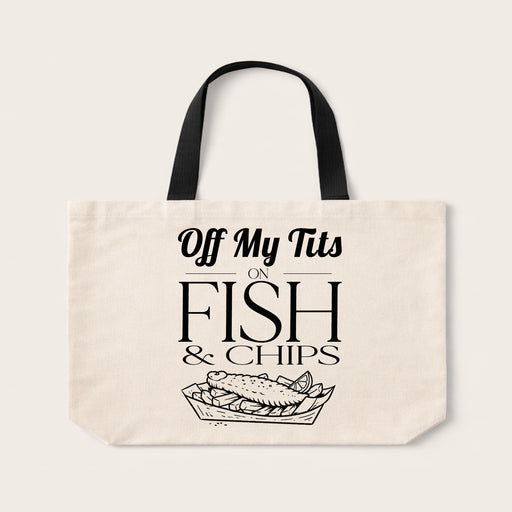 An off-white canvas tote bag with black handles and a black graphic print that reads 'Off My Tits ON FISH & CHIPS' above a fish in a tray illustration, shown on a light beige background.